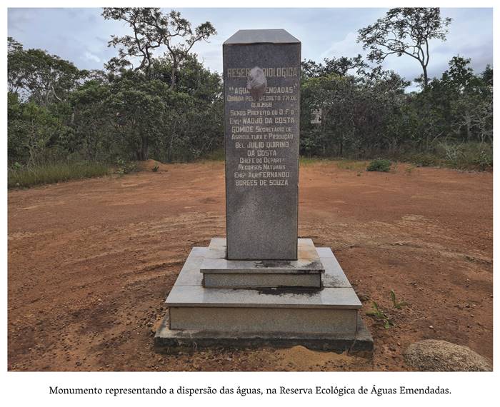   4 l- Monumento representando a dispersão, na reserva Ecológica de Águas Emendadas. O monumento está fixado numa base cinza clara, tem formato de pirulito na cor cinza escuro. No chão de terra batida. Ao fundo, um arvoredo e nesse monumento Lê-se o seguinte conteúdo. Reserva Biológica Águas Emendadas Criada pelo Decreto 771 de 12.8.1968 Sendo Prefeito do DF o Engenheiro Wadjô da Costa Gomide Secretário de Agricultura e Produção Bel. Júlio Quirino da Costa Chefe do Departamento de Recursos Naturais Eng. Agrônomo Fernando Borges de Souza. Realização: Academia Planaltinense de Letras, Artes e Ciências (APLAC), pelo Ecomuseu Pedra Fundamental e pelo Coletivo Nativo Audiodescrição produzida  pelo Instituto de Promoção das Pessoas com Deficiência Visual Audiodescritora: Elma Lúcia Rodrigues Consultor: Fernando Rodrigues Este projeto é promovido com recursos do Fundo de Apoio a Cultura do DF